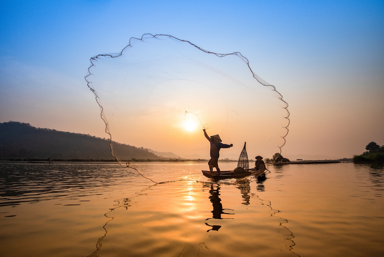 Asia fisherman net using on wooden boat casting net sunset or sunrise in the Mekong river / Silhouette fisherman boat with mountain background people life on countryside